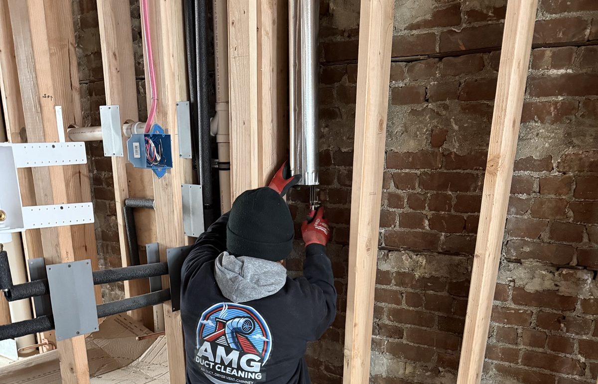 An AMG technician fitting a duct pipe between wall studs against an old brick wall