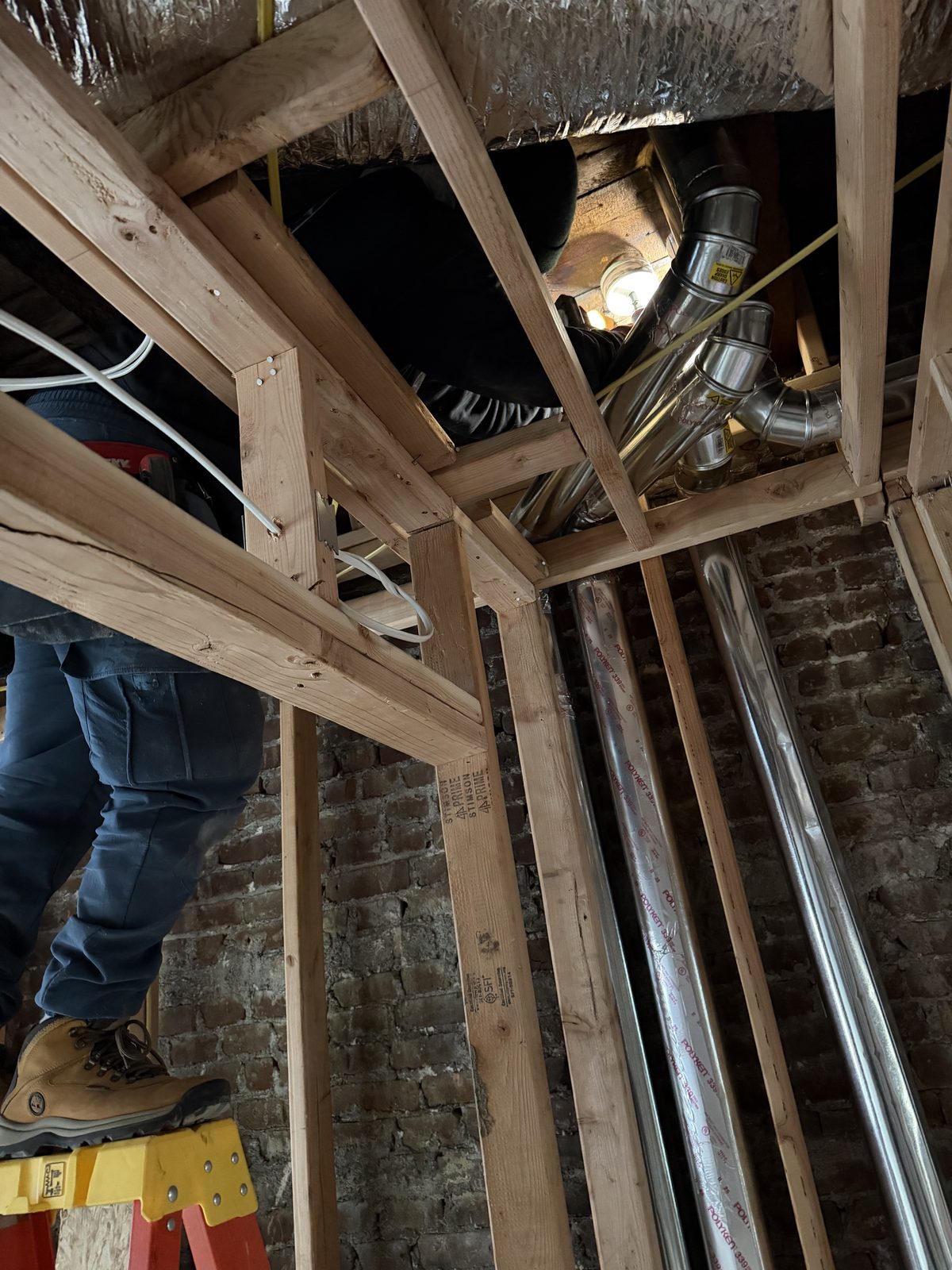 Round duct pipes run through open ceiling framing, a technician on the ladder above