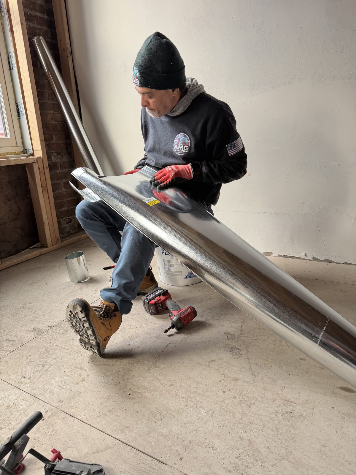 An AMG technician kneeling with a long section of galvanized duct pipe