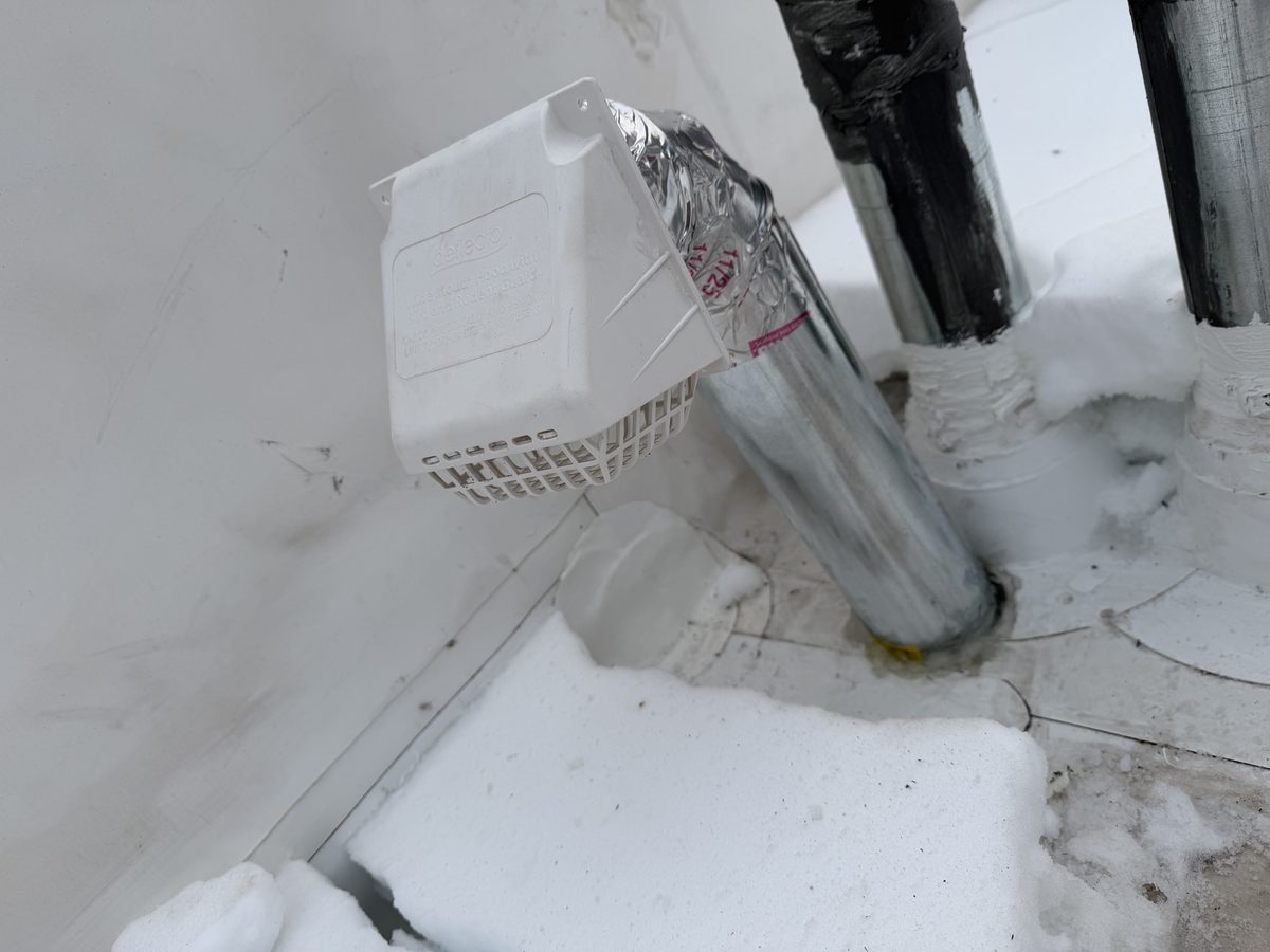 An exterior vent hood and its metal pipe at a snowy foundation