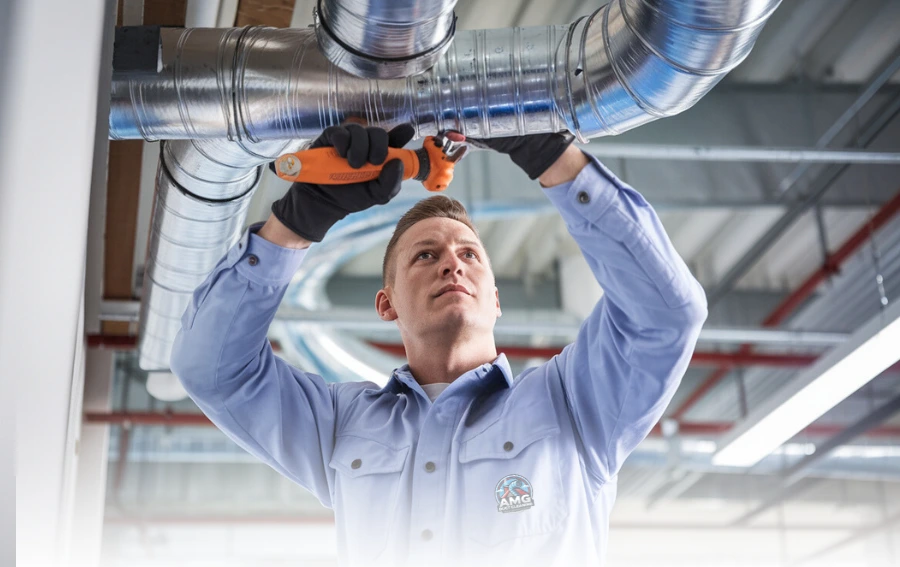 An HVAC technician wearing gloves and a uniform works on a duct system, using a wrench to adjust metal air ducts in an industrial or commercial setting.