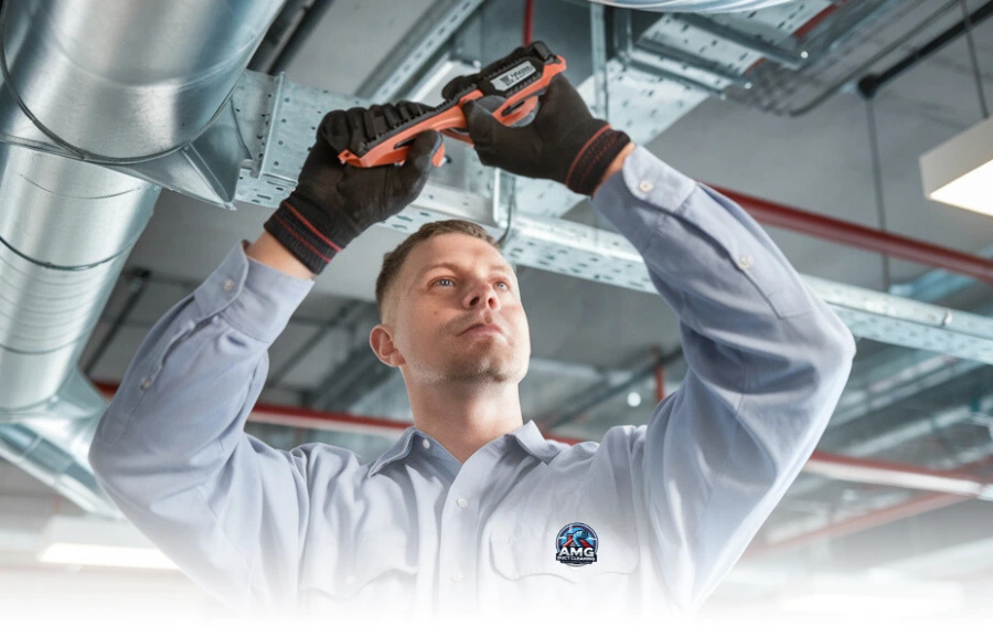 HVAC technician in a light blue uniform and black gloves using an adjustable wrench to work on an industrial air duct system in a commercial building with exposed metal beams and ventilation pipes.