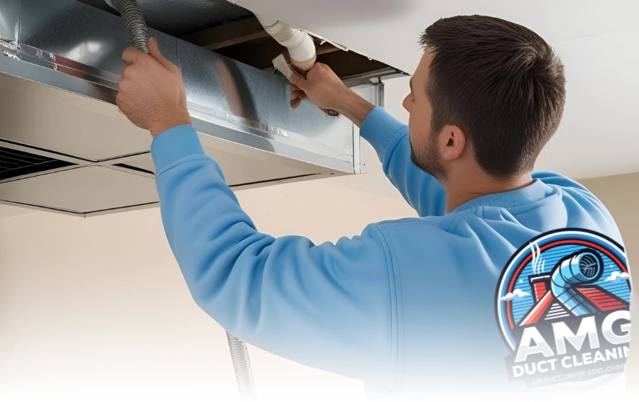 Technician wearing a light blue uniform inspecting and cleaning an air duct system in a residential ceiling, using a specialized vacuum hose. The "AMG Duct Cleaning" logo is prominently displayed in the bottom right corner.